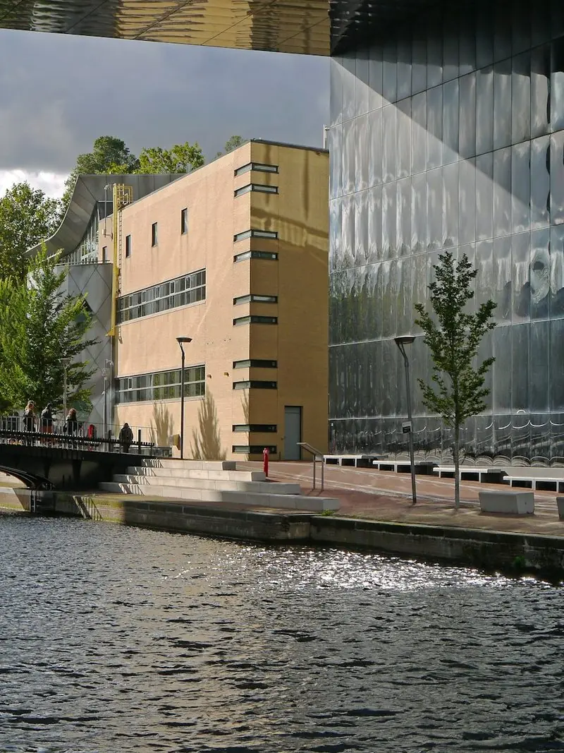 A modern glass and brick office building on the edge of an Amsterdam canal, seen from the bridge on an overcast day